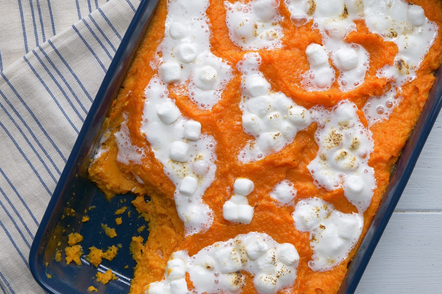 A casserole dish filled with sweet potato casserole is topped with melted marshmallows and sits on a striped cloth. Some portions of the casserole have been served, revealing an empty section of the dish. The bright orange color contrasts with the white of the marshmallows.