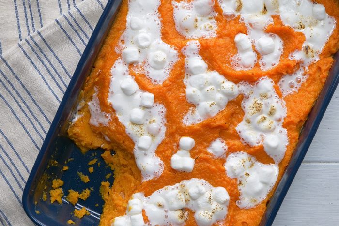 A casserole dish filled with sweet potato casserole is topped with melted marshmallows and sits on a striped cloth. Some portions of the casserole have been served, revealing an empty section of the dish. The bright orange color contrasts with the white of the marshmallows.
