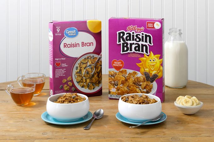 Kellogg's Raisin Bran and Walmart's Great Value Raisin Bran on a table with bananas, milk and tea