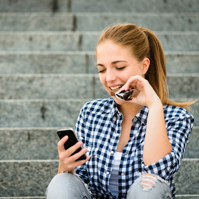 Teenager - young woman eating chocolate in street and looking in phone; Shutterstock ID 225964021; Job (TFH, TOH, RD, BNB, CWM, CM): TOH
