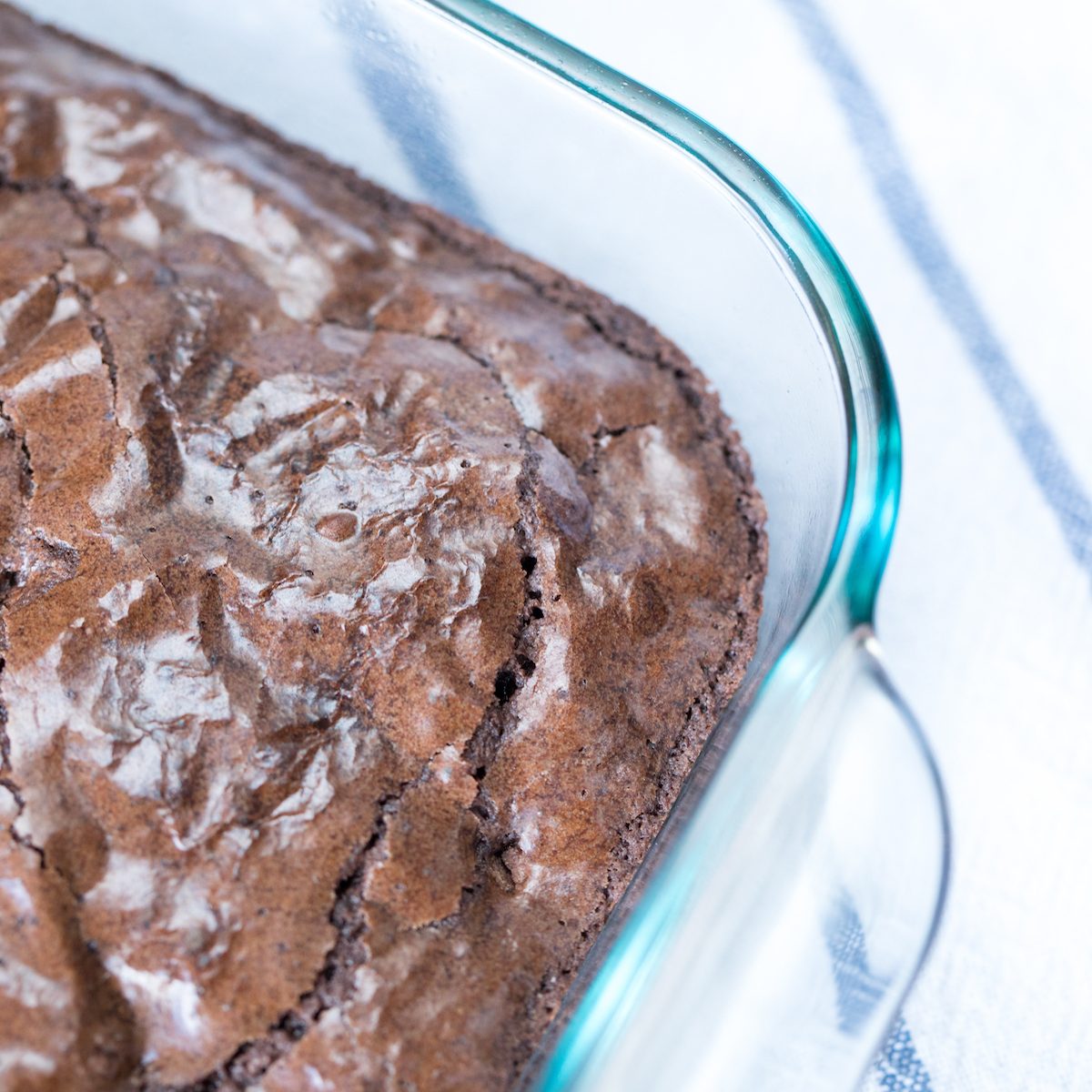 Overhead perspective on corner of cracked brown surface of freshly homemade baked chocolate chip chunk brownies in glass pan container on white and blue stripe linen table cloth in soft window light