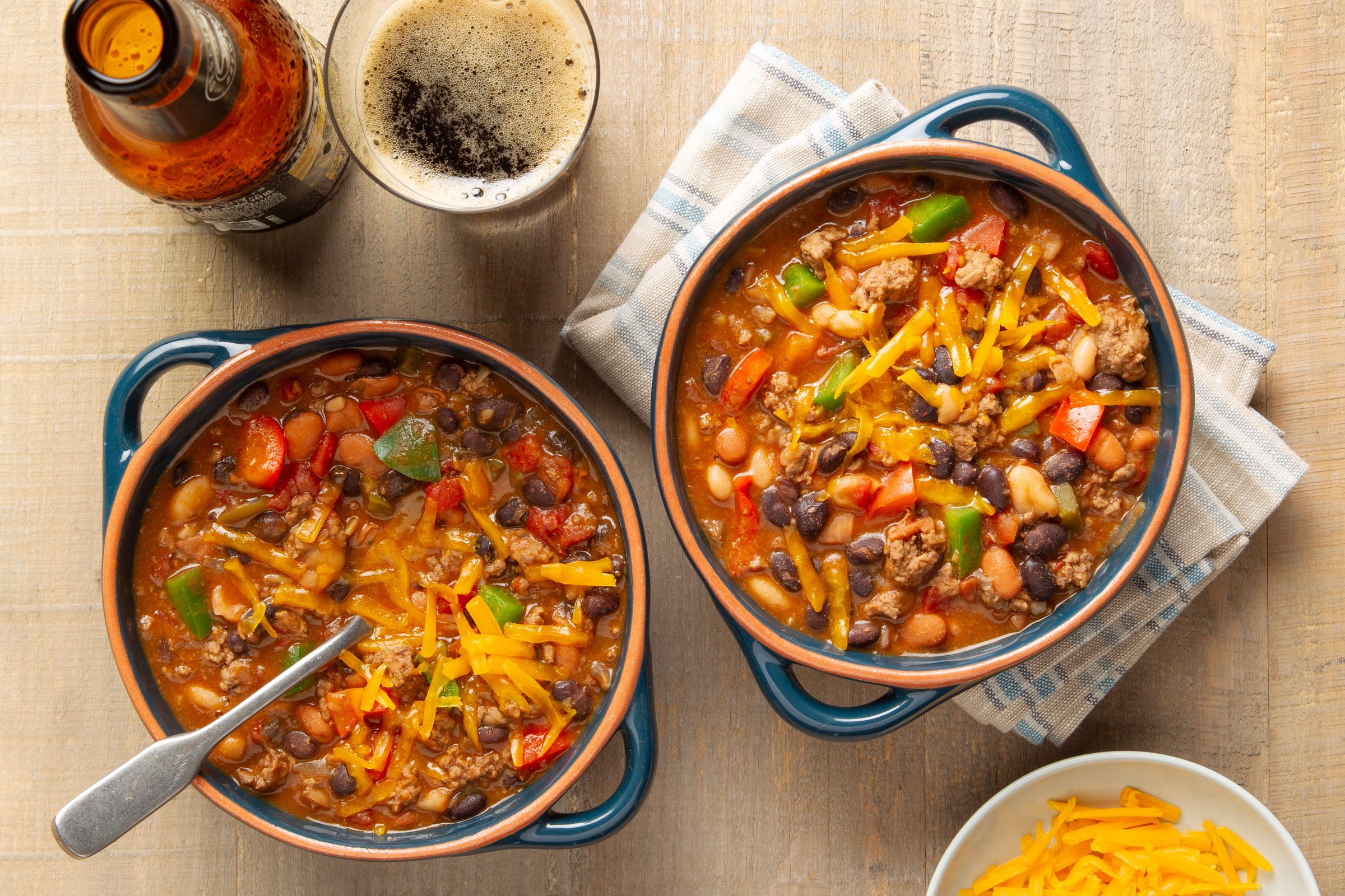 two crocks of chili on a wooden table; bottle and glass of beer nearby