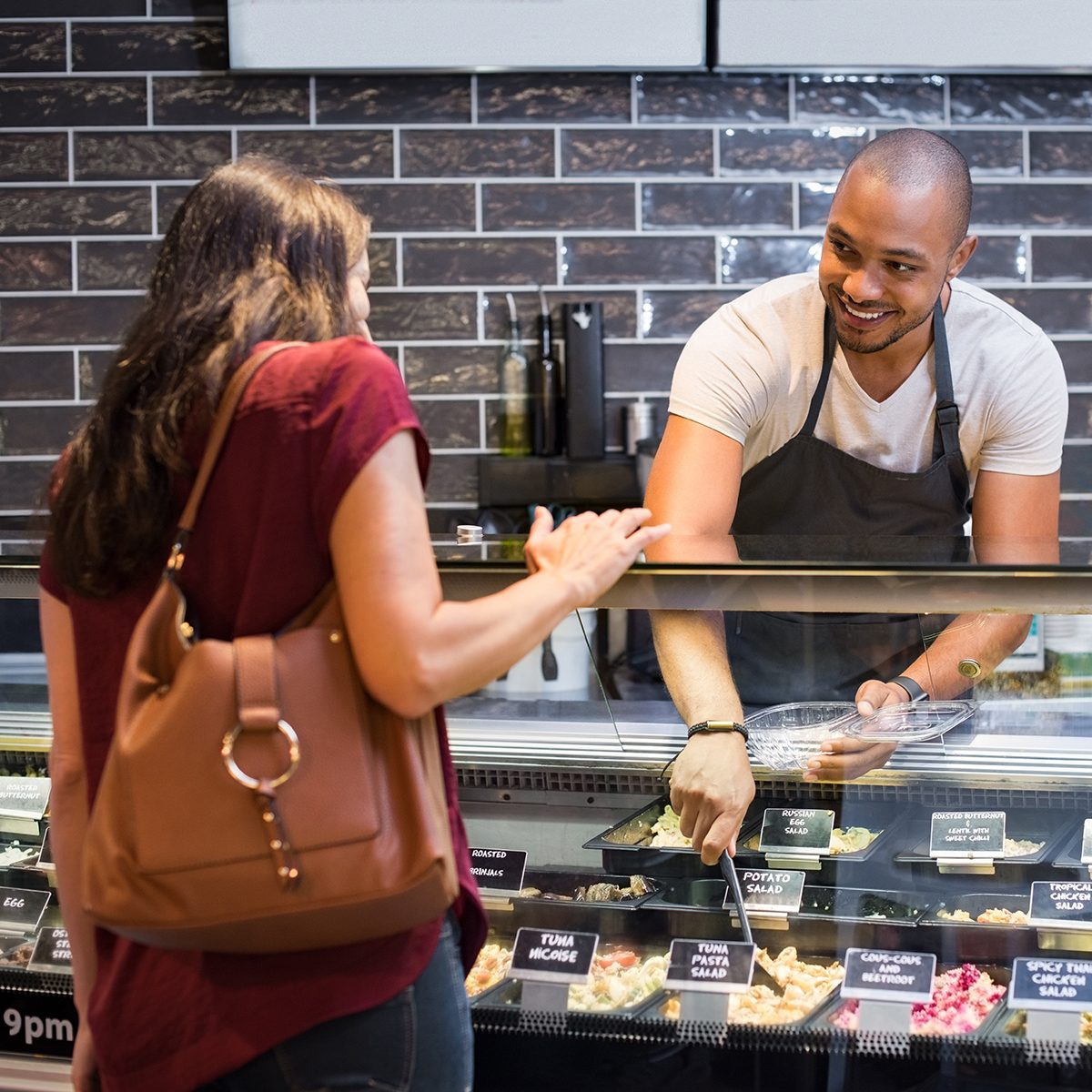 African waiter serving fresh food to young woman. Happy smiling guy preparing take away salad for woman customer. Black man with apron taking pasta salad in spoon and serving to customer.