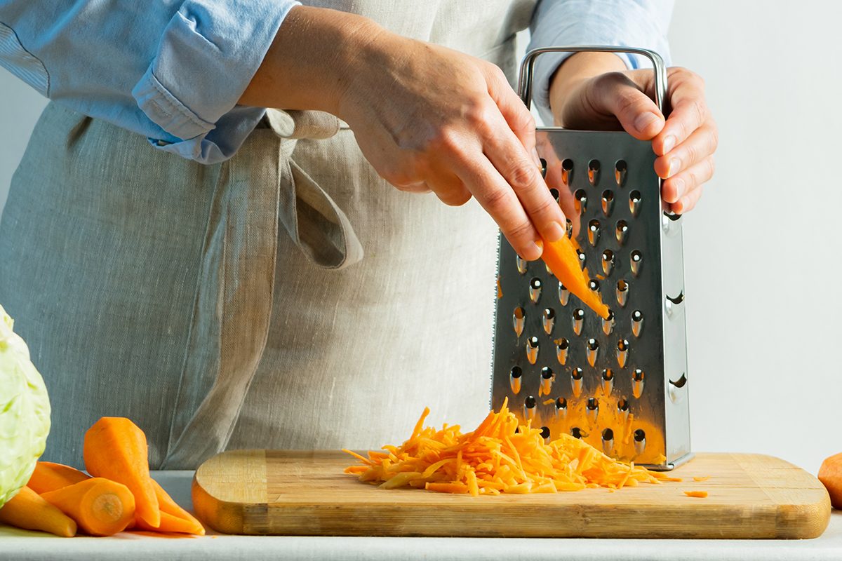 Woman Grates Fresh Carrots For Cooking Salad Or For Salting Cabbage Natural Background. Vegetarian Food Concept.