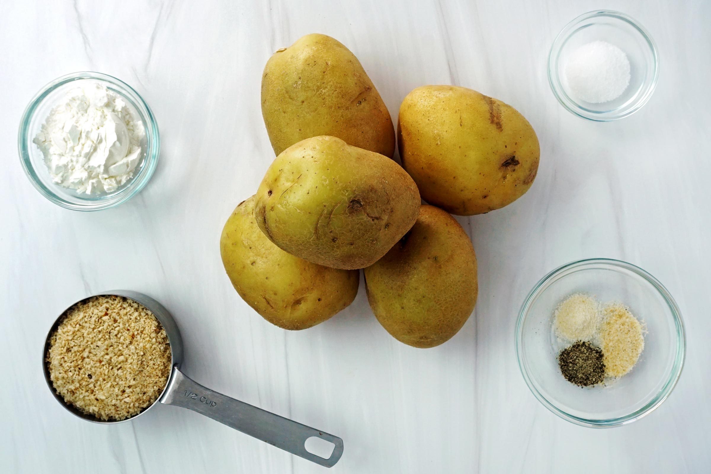 a pile of potatoes surrounded by small bowls and measuring cups filled with other ingredients on a white table cloth, view from above