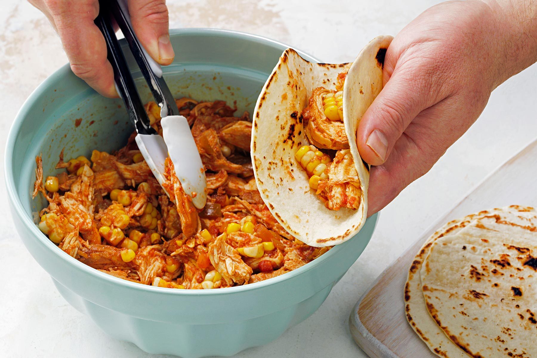 Picking up shredded chicken mixture with tongs; taco shell in one hand