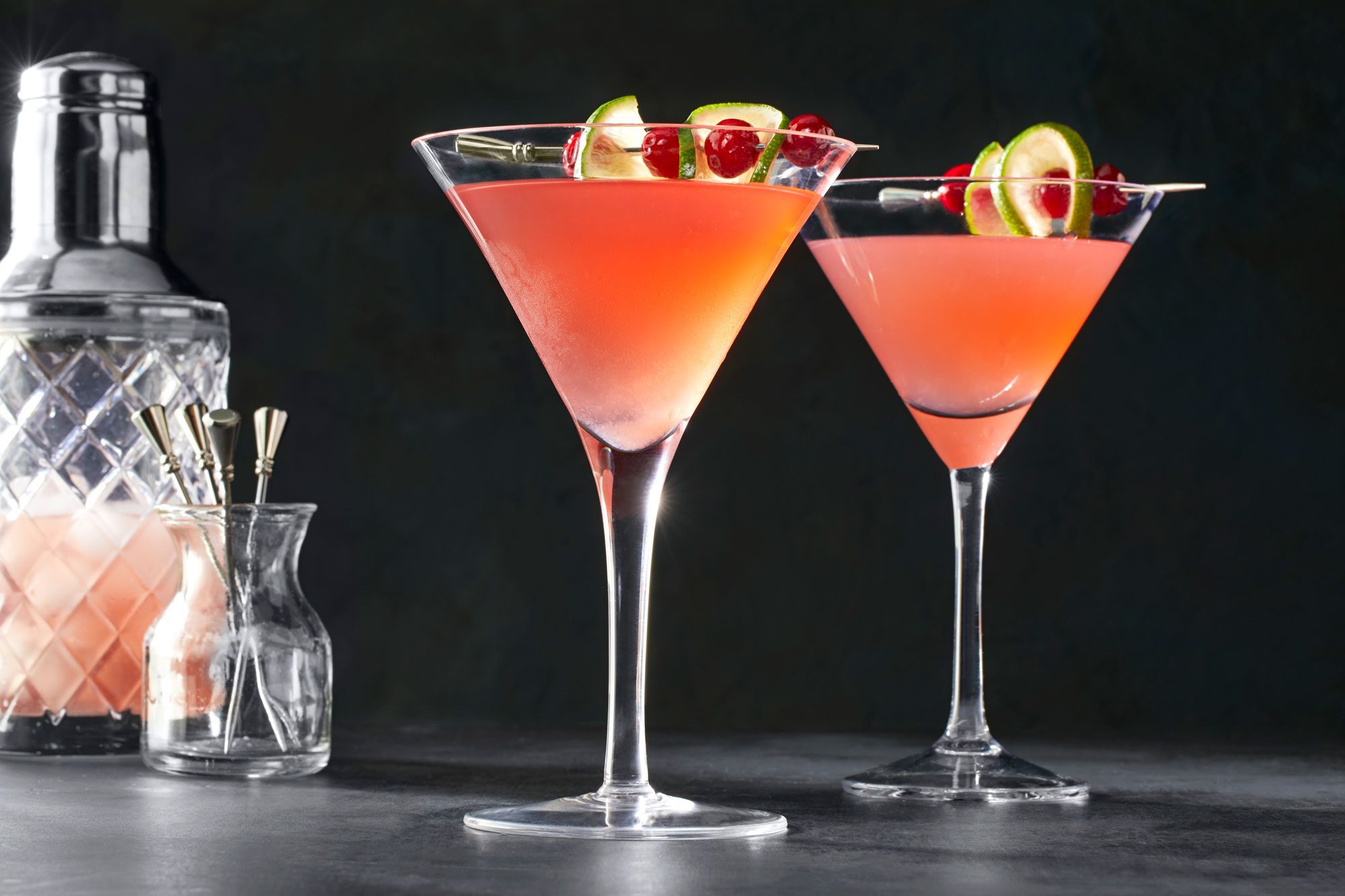 Cosmopolitan in cocktail glasses garnished with lime and cranberries with shaker on the side