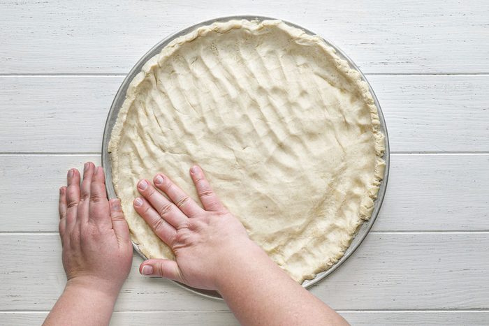 pressing the pizza crust dough on a pizza pan on white painted wooden surface