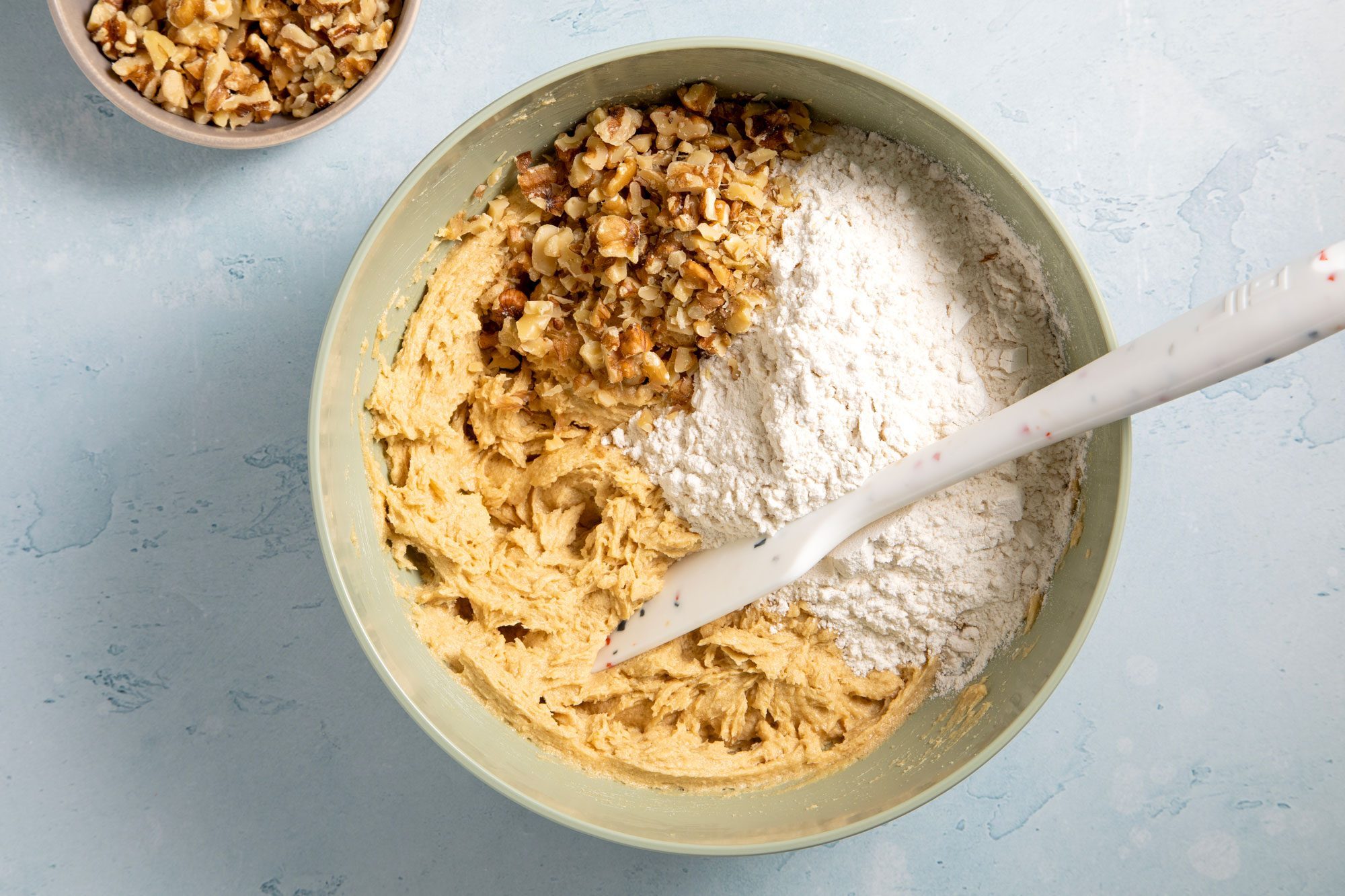 overhead shot; light blue textured background; added flour and walnuts in beaten mixture in a large bowl;