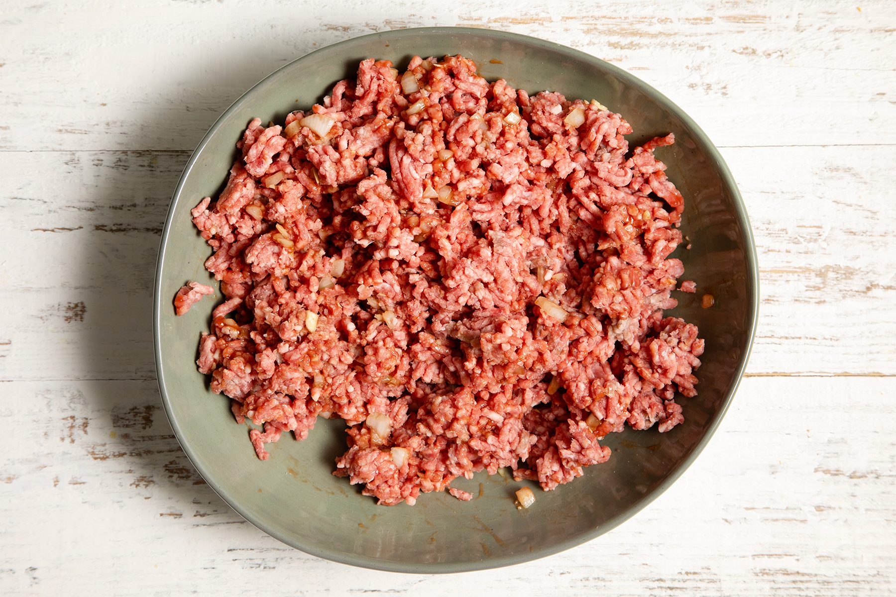 A large oval bowl filled with ground meat sits on a rustic white wooden surface.