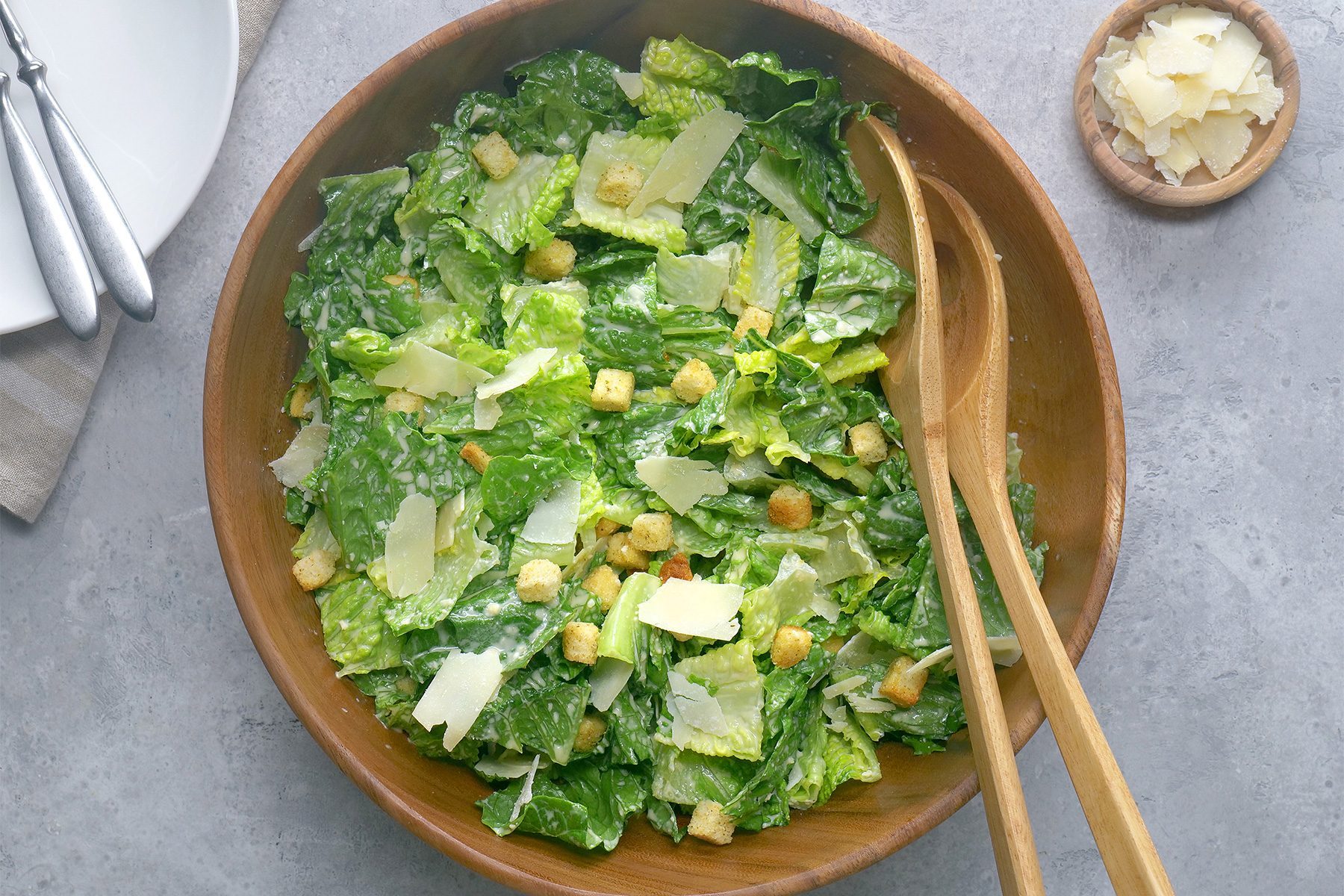 Overhead shot of Ceasar Salad; in a large bowl; sprinkle with croutons and parmesan; wooden spoon; white plate; cutlery; napkin; grey background