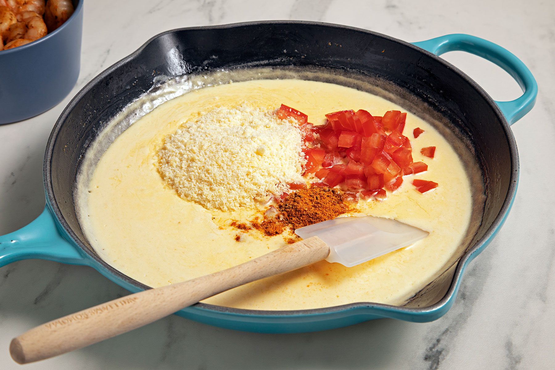 A blue skillet on a marble surface with a creamy yellow mixture being prepared. The mixture contains a pile of shredded cheese, chopped tomatoes, and spices. A white spatula with a wooden handle rests in the skillet.