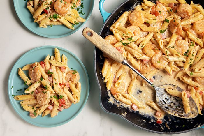 A skillet filled with creamy shrimp pasta sits next to two blue plates, each with a serving of the pasta. The dish includes penne pasta, shrimp, and chopped tomatoes, garnished with fresh herbs. A slotted spoon rests in the skillet.