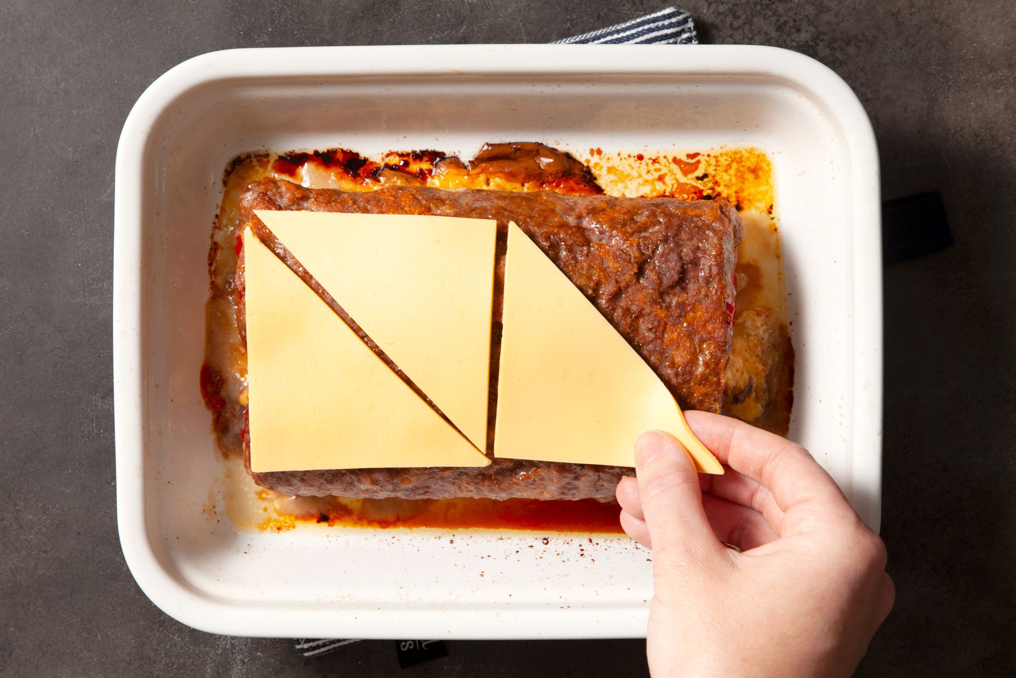Overhead shot of cut the reserved cheese slices in half diagonally; place on top of loaf; baking pan; white and black line base cloth; black texture background;