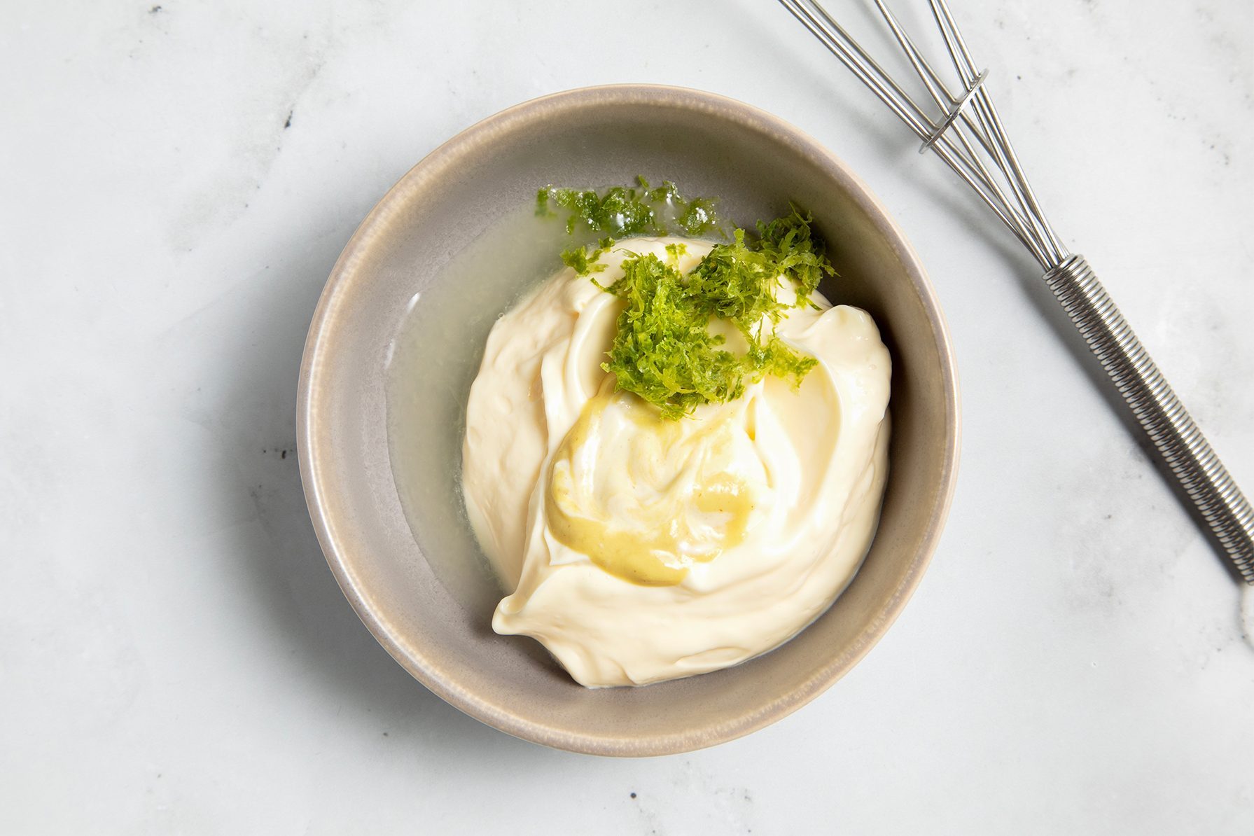 A beige bowl filled with creamy mayonnaise garnished with a sprinkling of lime zest on top sits on a white marble surface.