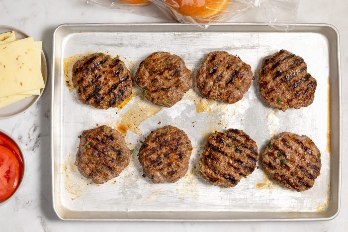 A baking tray with eight grilled beef burger patties, positioned in two rows.