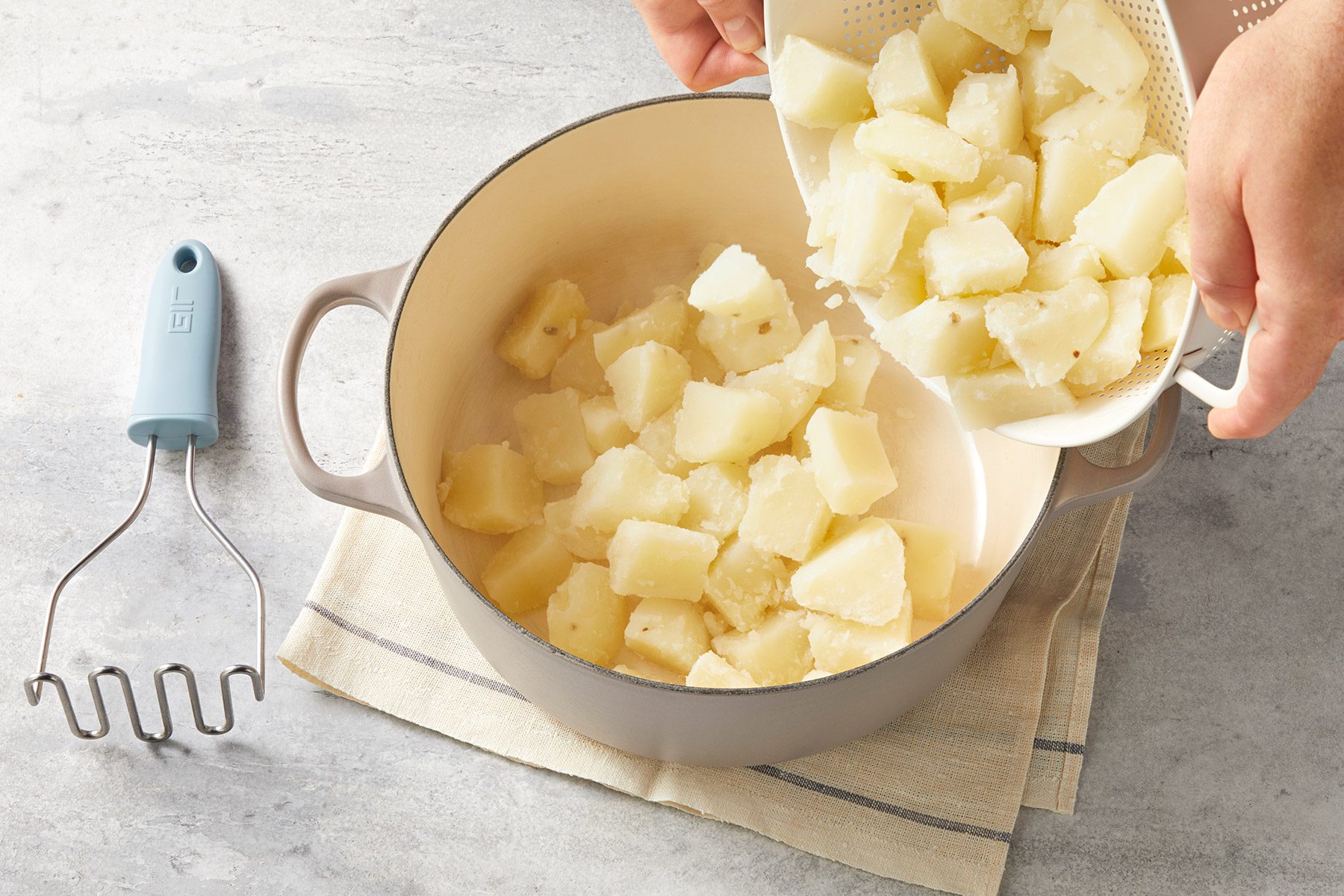 Hands are seen pouring cooked, cubed potatoes from a strainer into a beige pot on a gray surface. A potato masher with a blue handle lies to the side on a light-colored folded cloth.