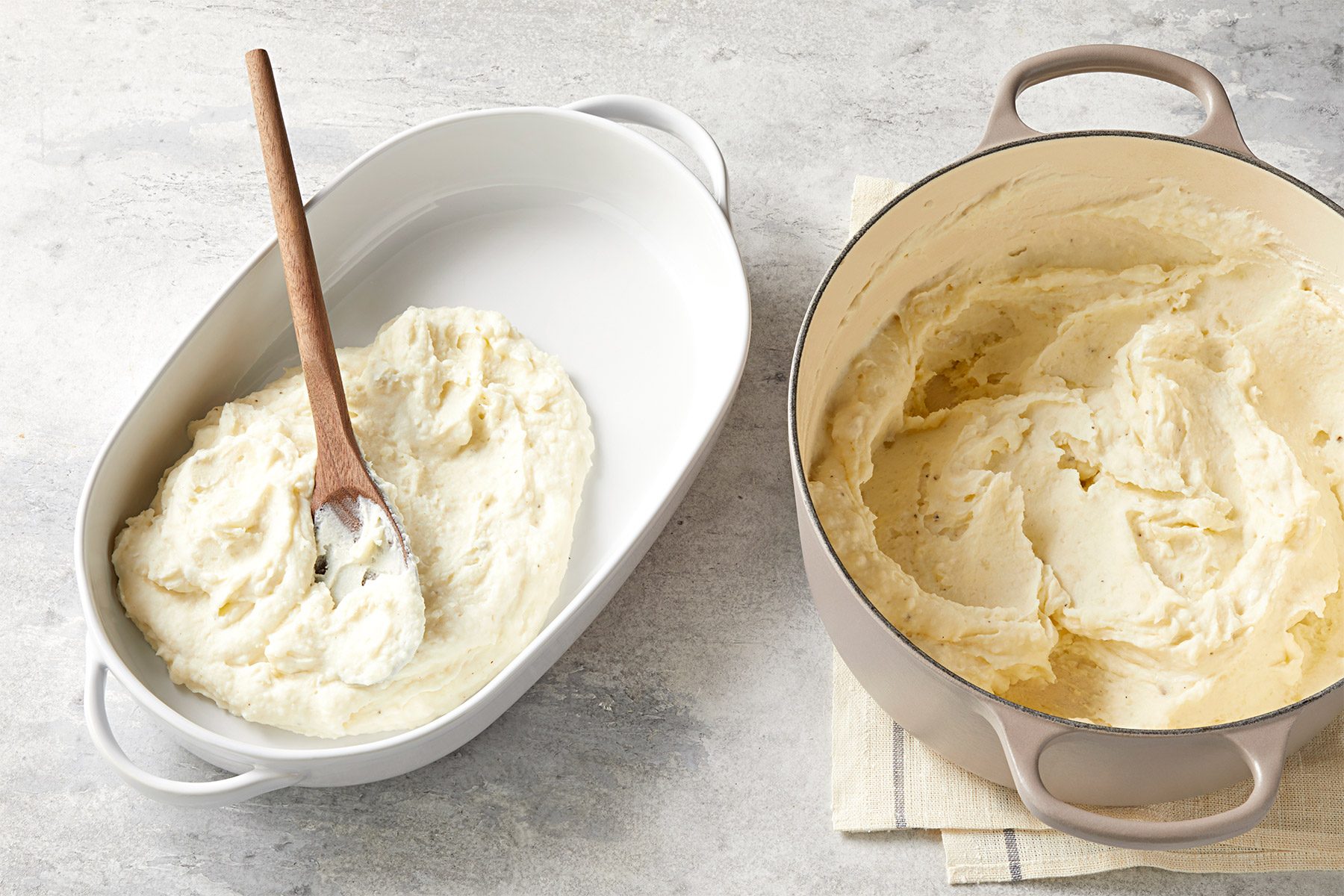 A wooden spoon rests in a white ceramic baking dish filled with mashed potatoes. Next to it is a dutch oven containing more mashed potatoes on top of a folded cloth, with both items placed on a light-colored stone surface.