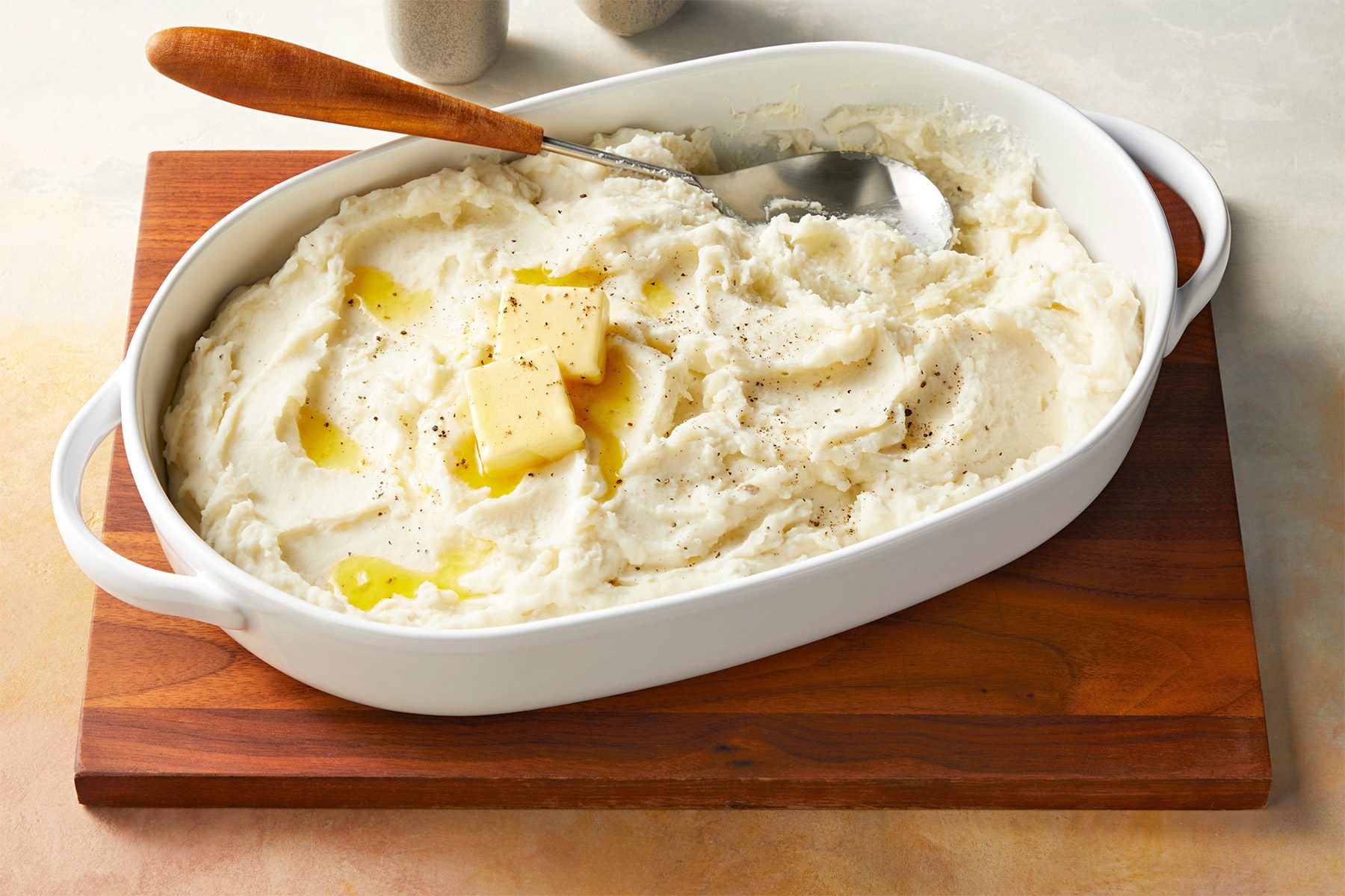 A white ceramic dish filled with creamy mashed potatoes, topped with two pats of melting butter. A wooden spoon is placed on the edge of the dish which is resting on a wooden board.