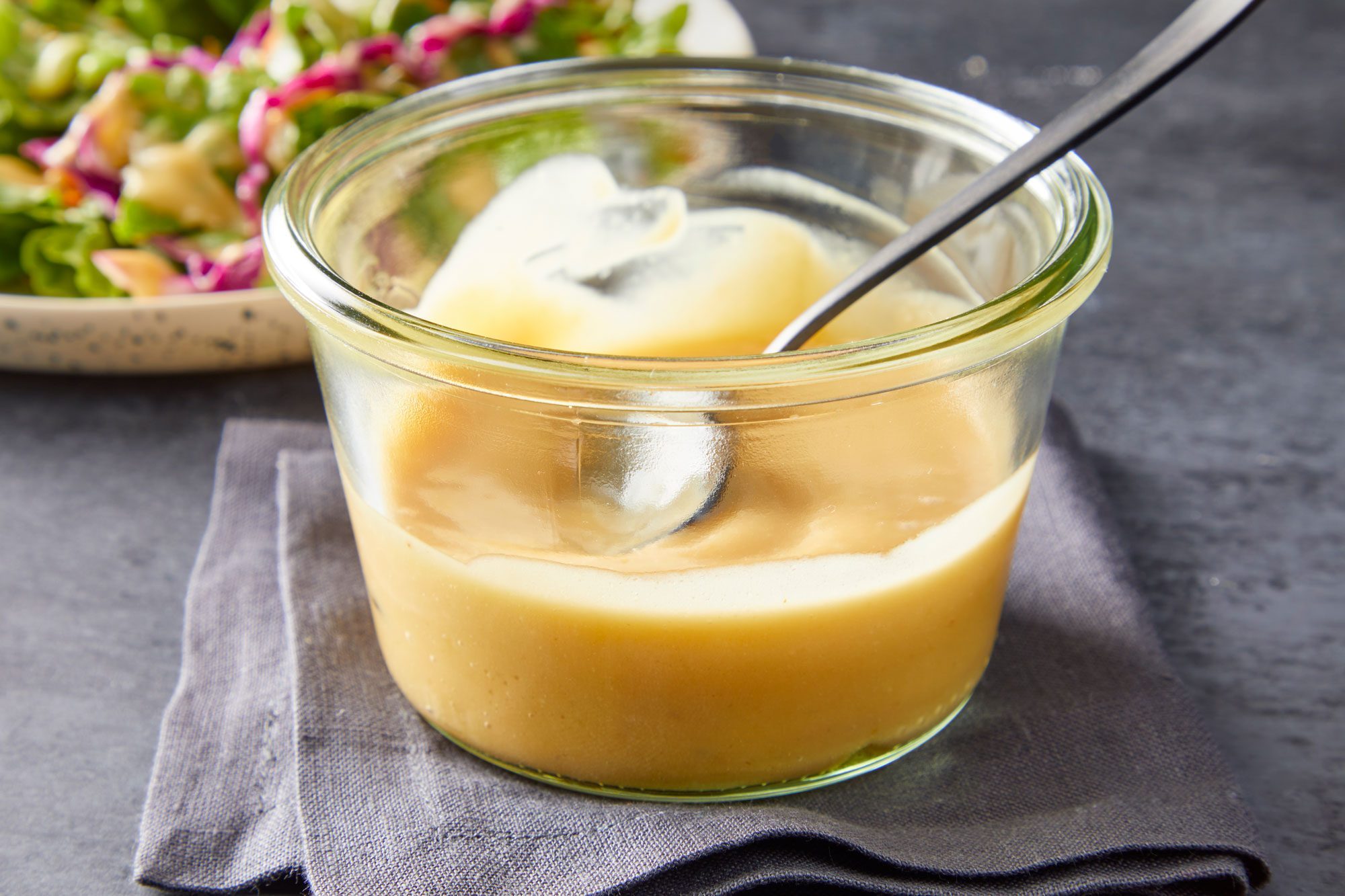Table view shot of Miso Dressing; in a glass bowl; pour over salad served on plate; spoon; napkin; grey background;