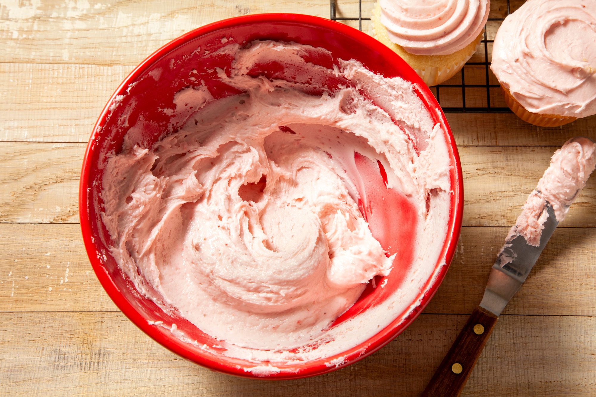 overhead shot of strawberry frosting in large red bowl and knife placed on right side and cup cakes on wired rack on wooden background