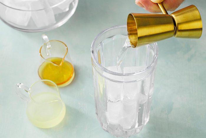 A hand holding a golden jigger is pouring a clear liquid into a tall glass filled with ice cubes. Nearby, two small pitchers containing yellow and green liquids are placed on a light blue surface.