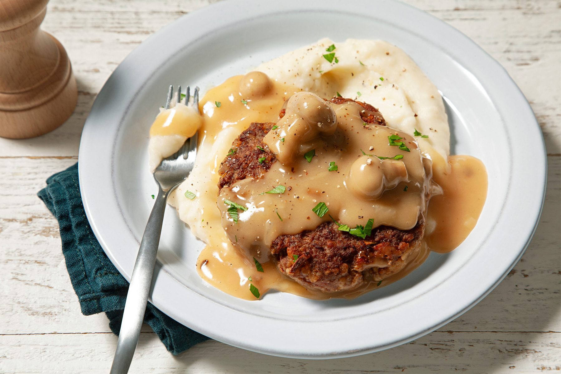 3/4th shot; wooden background; Hamburger Steaks with Mushroom Gravy served with mashed potatoes in a white plate with silver spoon over green kitchen towel;