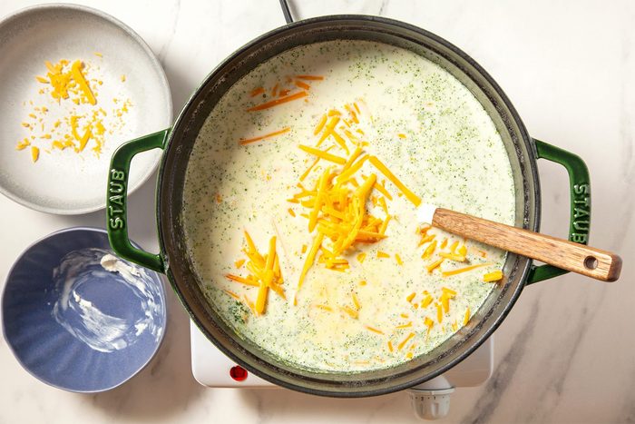 An overhead shot of a pot on a stovetop containing creamy broccoli cheese soup with shredded cheddar cheese on top and a wooden spoon resting inside. To the side are two bowls, one with remnants of sauce and the other with more shredded cheddar cheese.