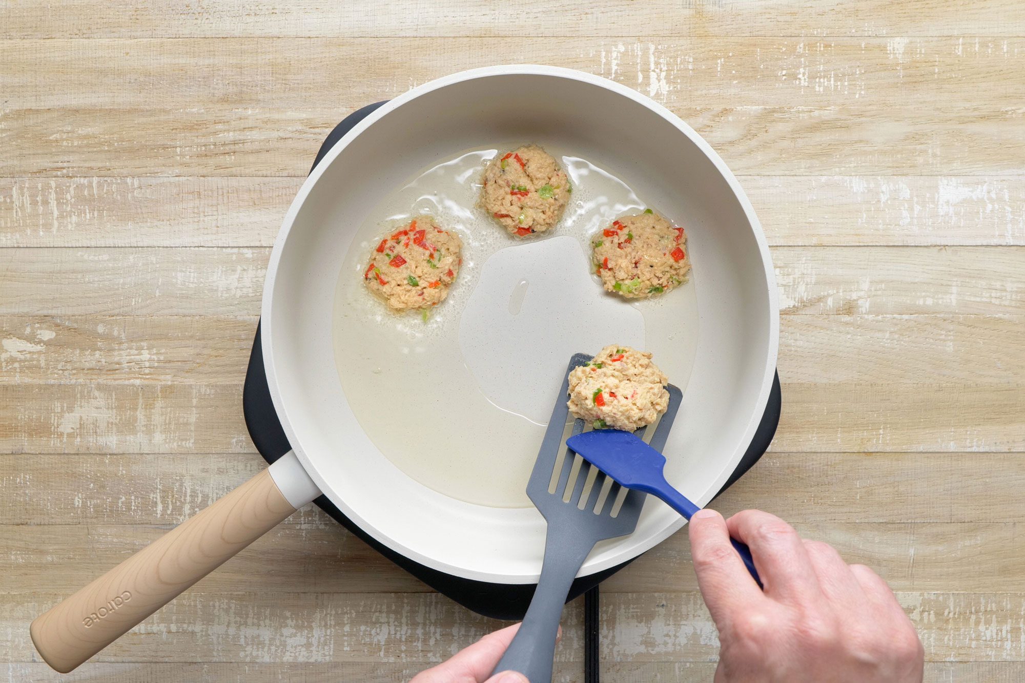 Overheda shot of a large skillet; heat oil over medium heat; add croquettes in batches; cook until golden brown; 3-4 mintues on each side; spatula burger flipper turner; small spatula; induction; wooden background;