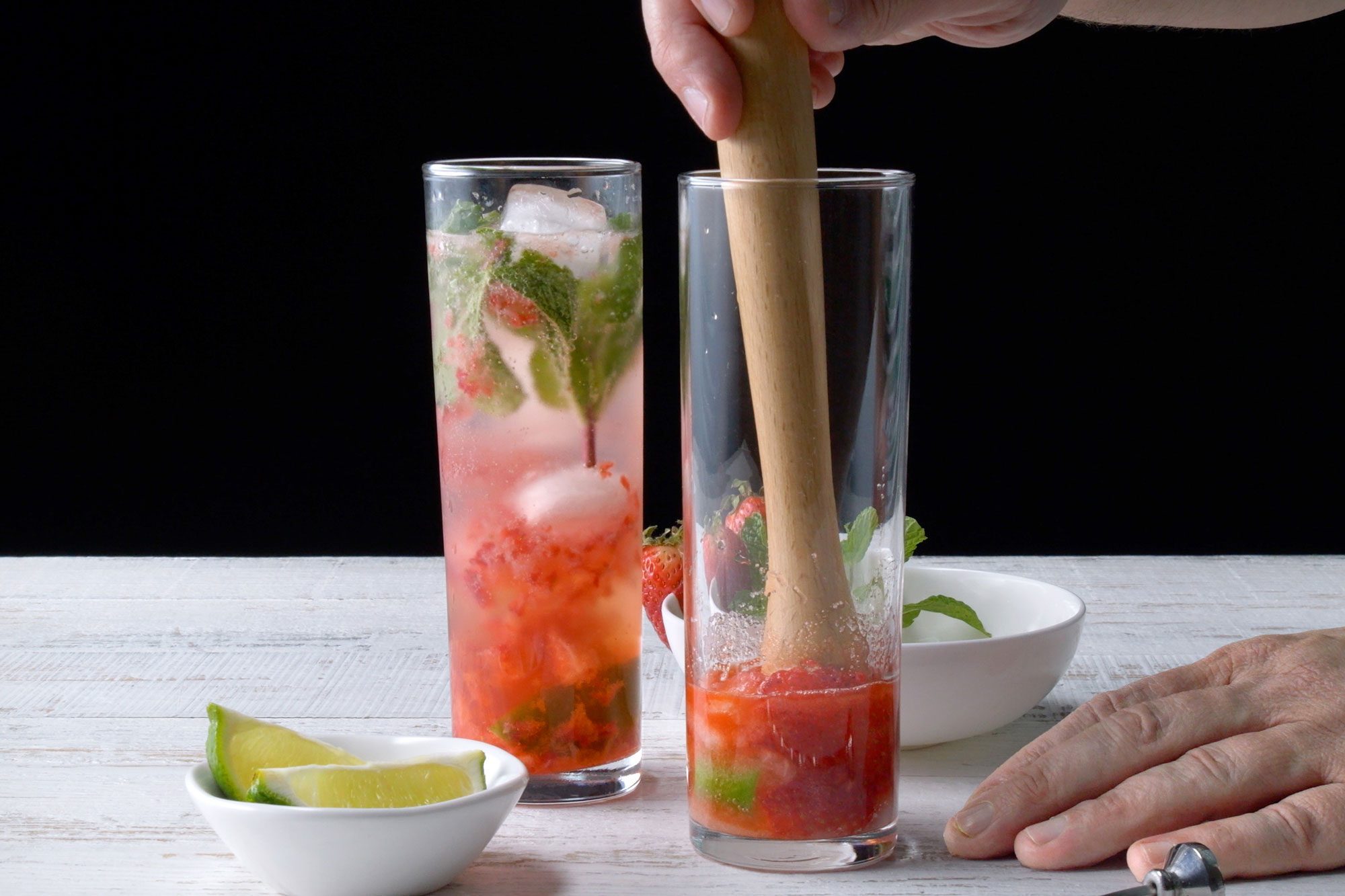 horizontal shot; black background; gently pressing ingredients with long wooden stick inside glass with hands;