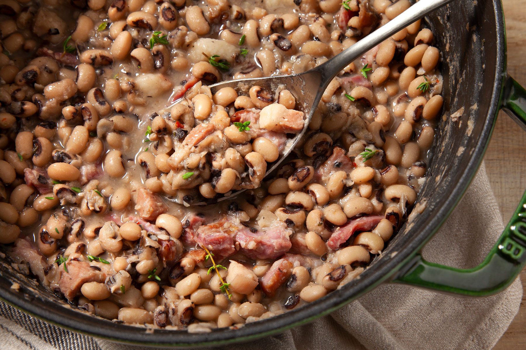 A pot of cooked black-eyed peas, mixed with chunks of ham and garnished with fresh thyme, sits on a beige cloth. A spoon is lifting a portion of the beans.
