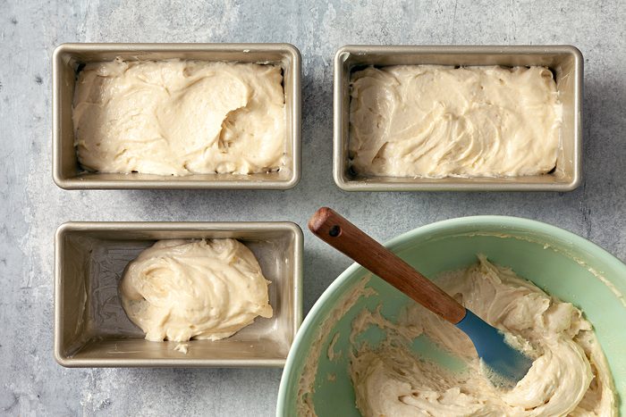 A bowl with banana bread batter and a rubber spatula on the right. On the left, batter has been poured into three rectangular loaf pans, two of which are full and one partially filled. The scene is set on a gray countertop.