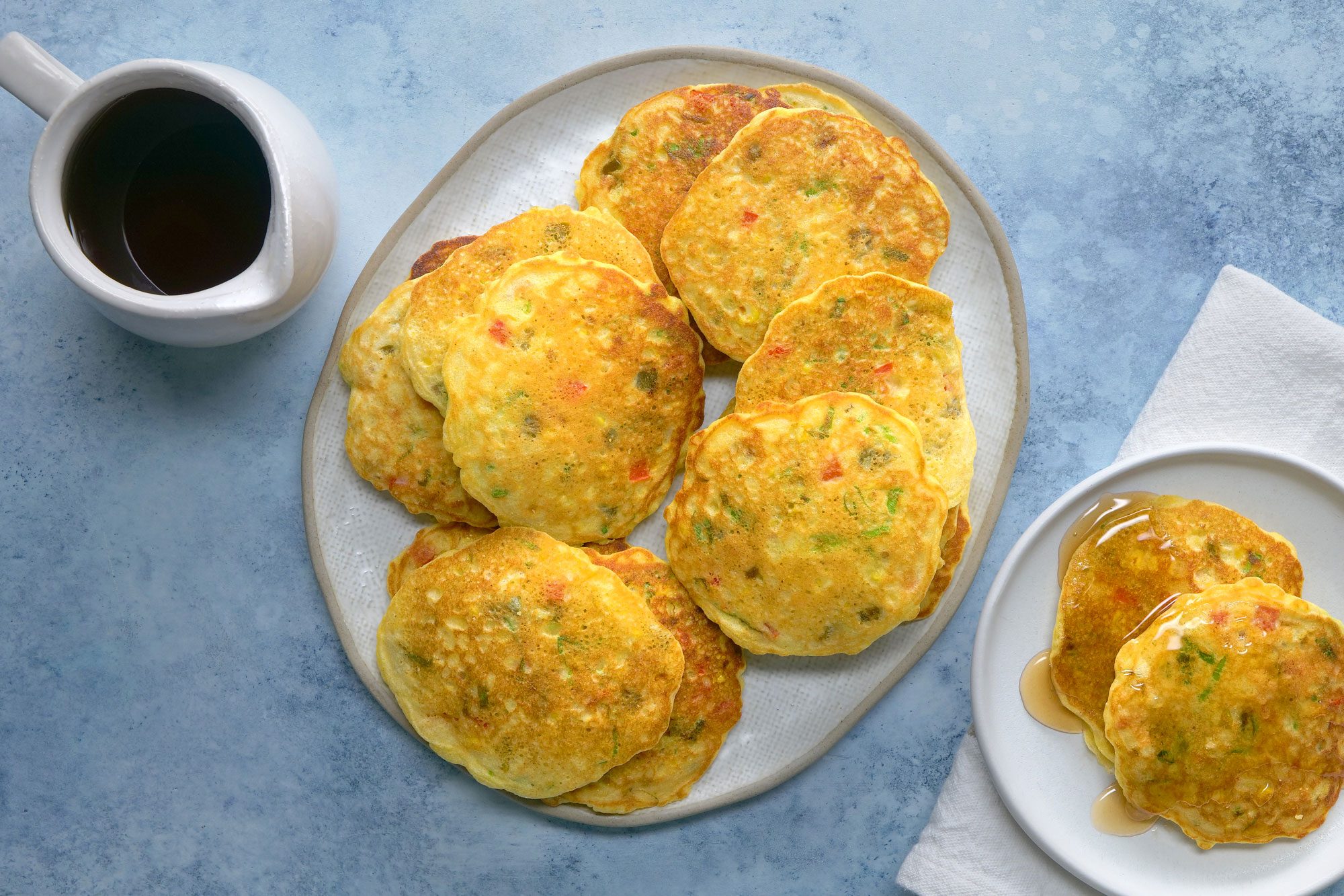 overhead shot; light blue background; Corn Cakes on a large white plate; Corn Cakes on small white plate over white napkin; honey in a jar;
