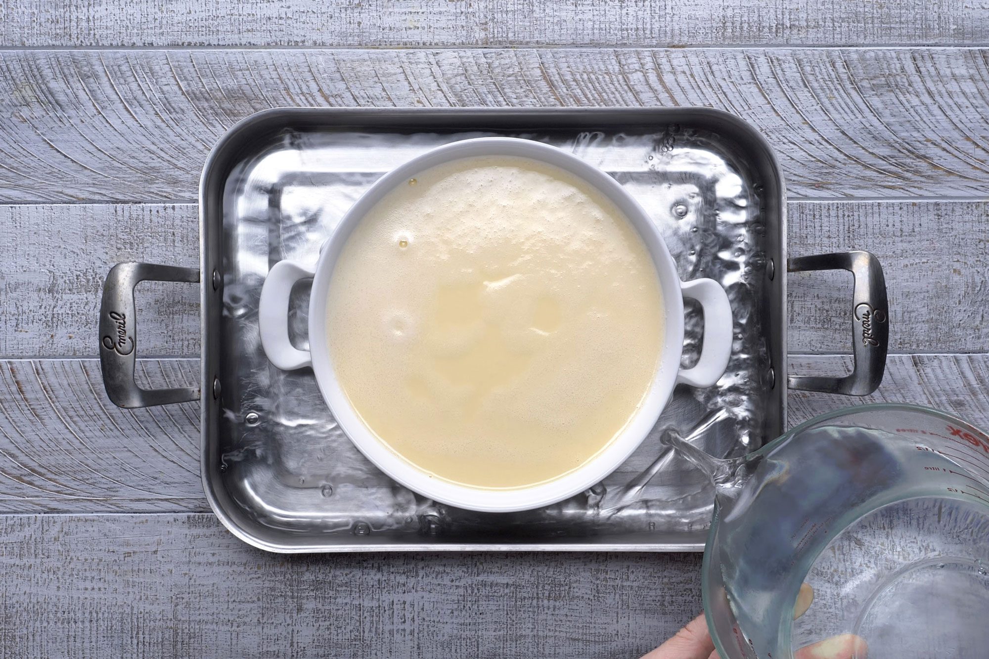 Overhead shot of place round baking dish in a larger baking pan; place pan on oven rack; add very hot water to pan to within 1/2 inch of top of round baking dish; bake until a knife inserted near the center comes out clean; wooden background;