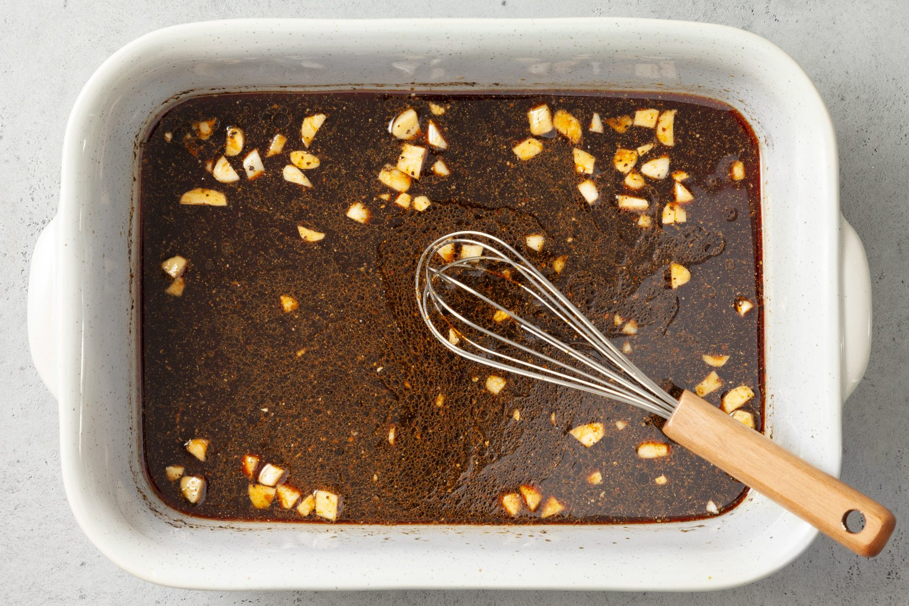 Top view of a baking dish filled with a marinade mixed with chopped garlic pieces. A whisk is resting in the dish.