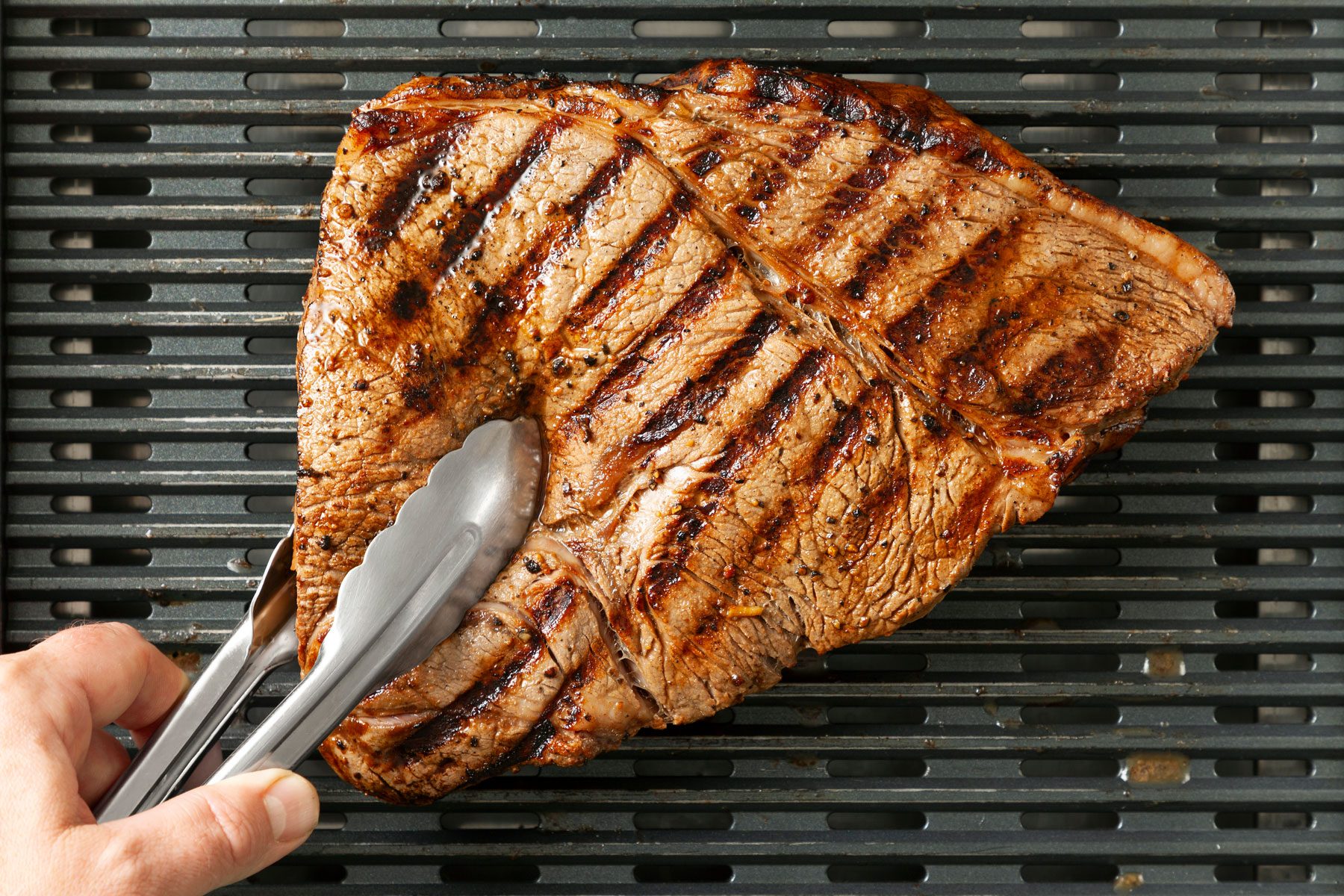 A large, perfectly grilled steak with visible char marks sits on a barbecue grill. A hand holding a pair of tongs is positioned at one end.