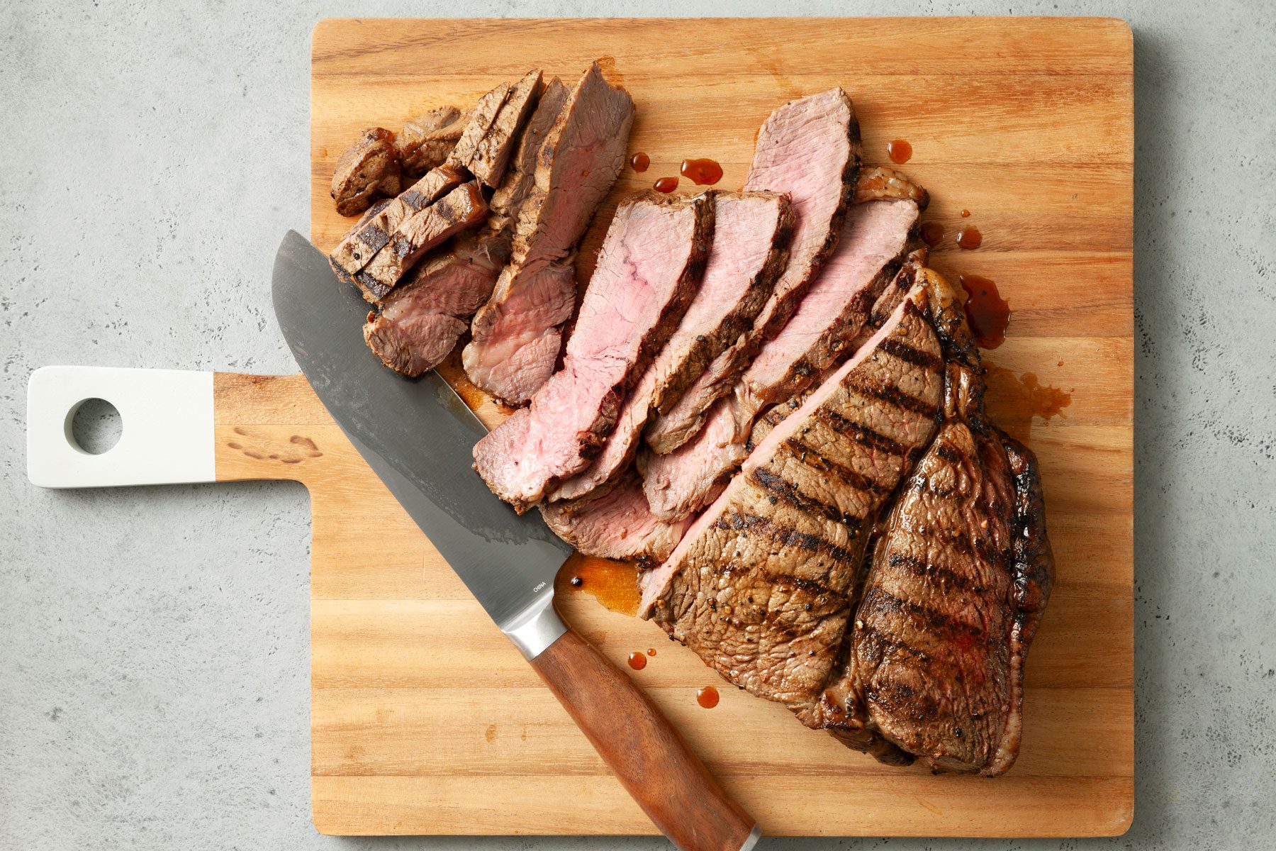 A wooden cutting board with several slices of medium-rare grilled steak. A large kitchen knife rests beside the steak.