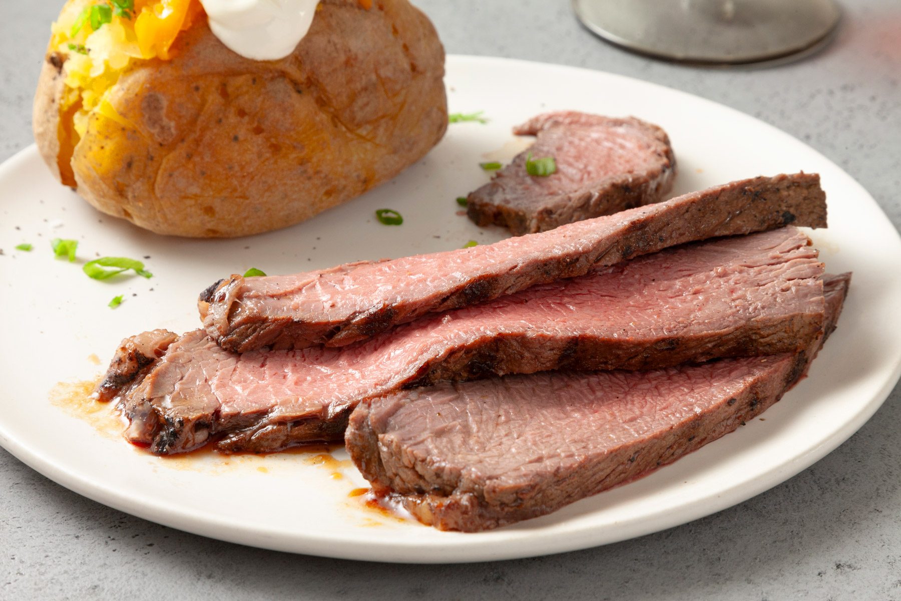 A white plate with slices of roast beef, garnished with chopped green onions. A baked potato topped with sour cream and melted cheese is in the background.