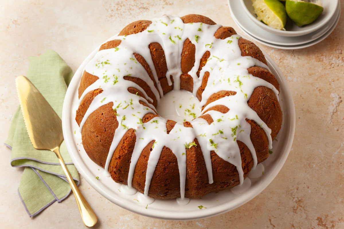 Full yield of Taste of Home Margarita Cake with a gold pie server on a green linen, stack of plates, squeezed limes in a bowl