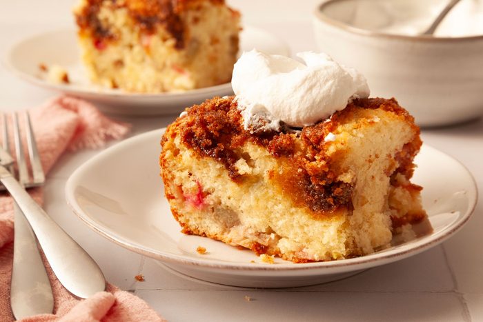 2 Single servings of Taste of Home Rhubarb Coffee Cake on white dessert plates with pink linen and a bowl of whipped cream