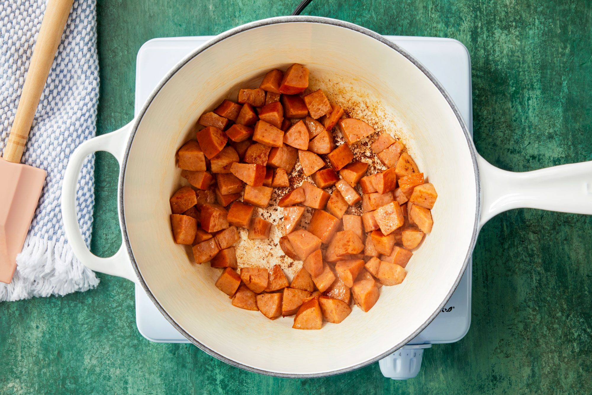 Overhead shot of a large saucepan; cook kielbasa over medium heat until lightly browned; drain and set aside; induction; spatula; kitchen towel; green texture surface;