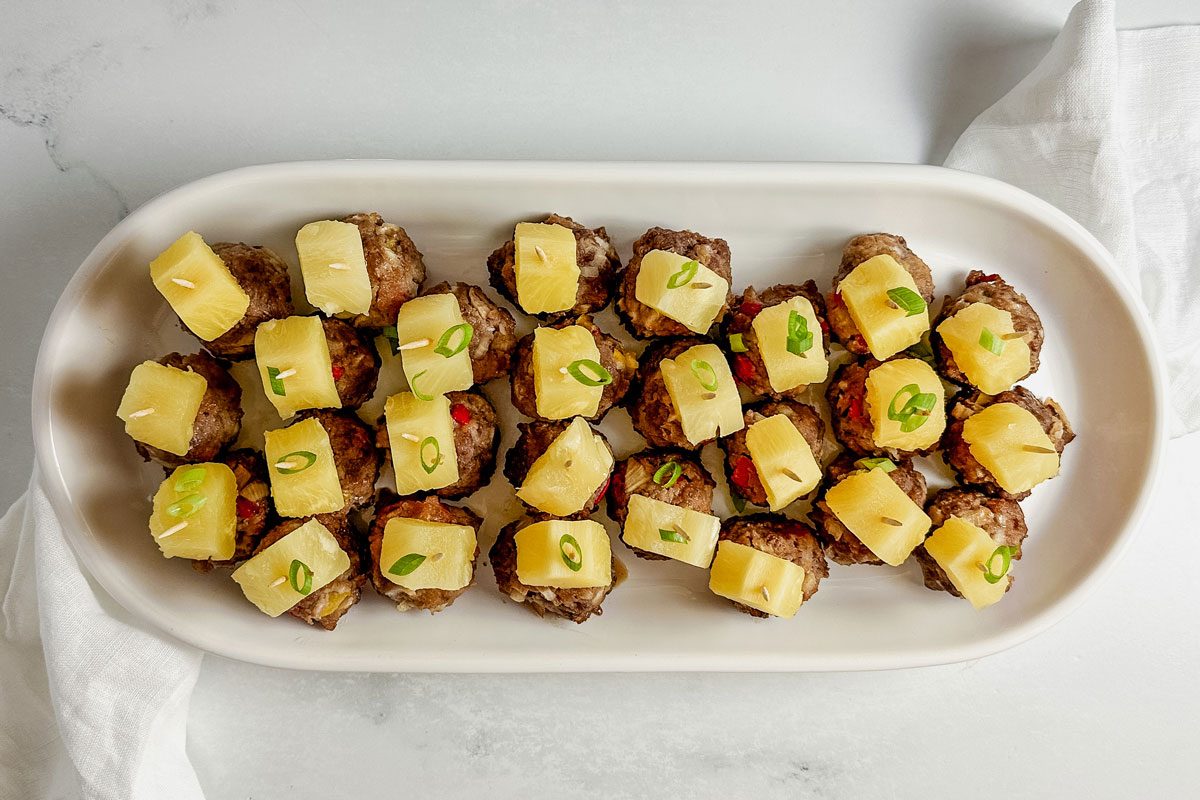 Taste of Home teriyaki meatballs topped with pineapple chunks in a white ceramic tray on a linen napkin on a marble surface.
