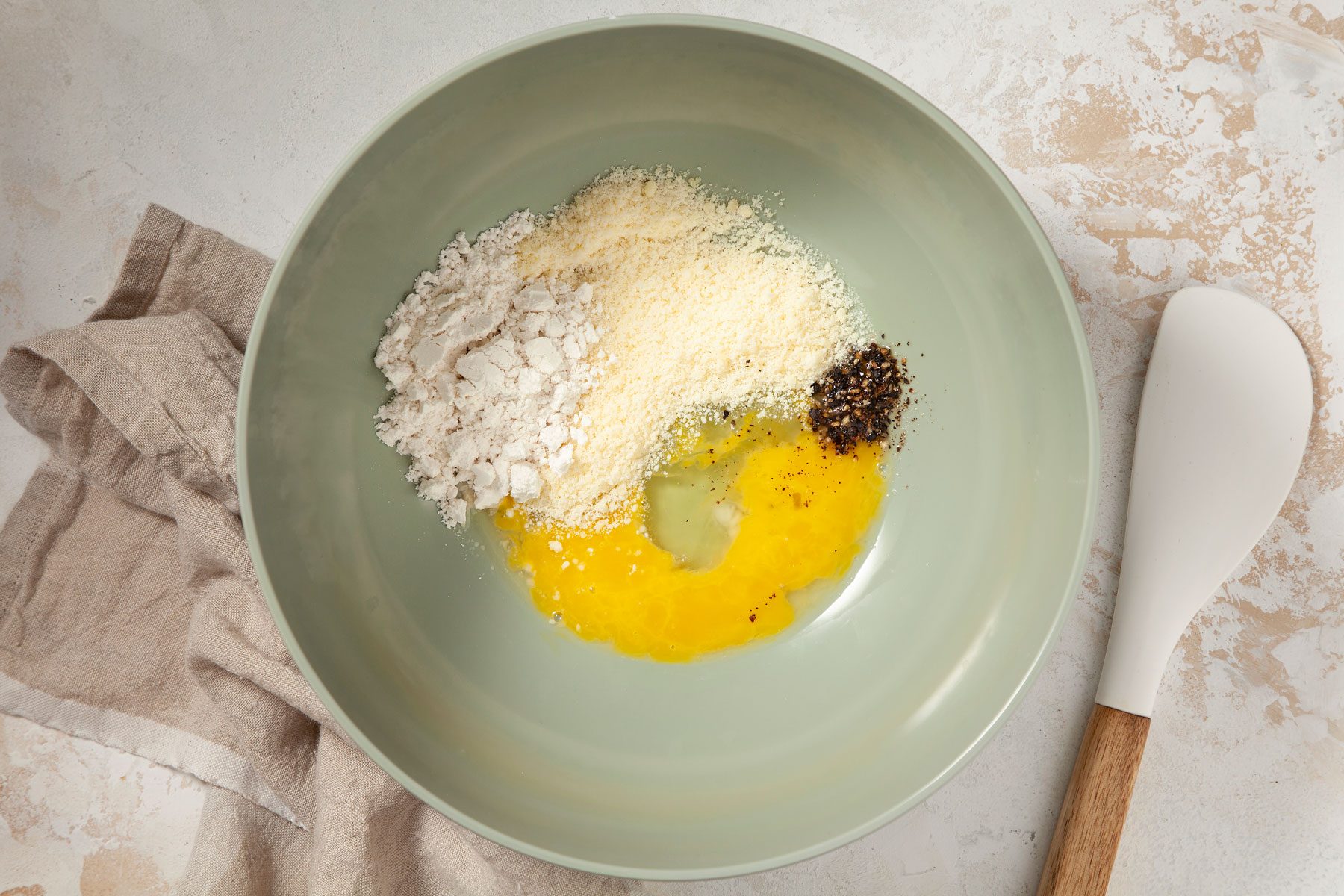  Egg, cheese, baking mix and pepper are in a bowl. A white spatula is by the side of the bowl.