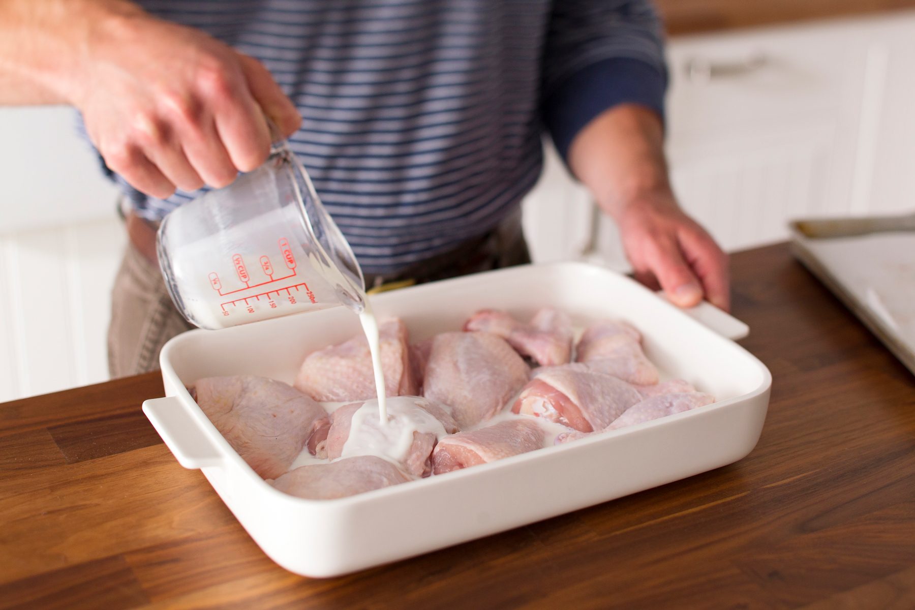 Raw chicken parts in a flat dish with high sides having buttermilk poured over them from a measuring cup