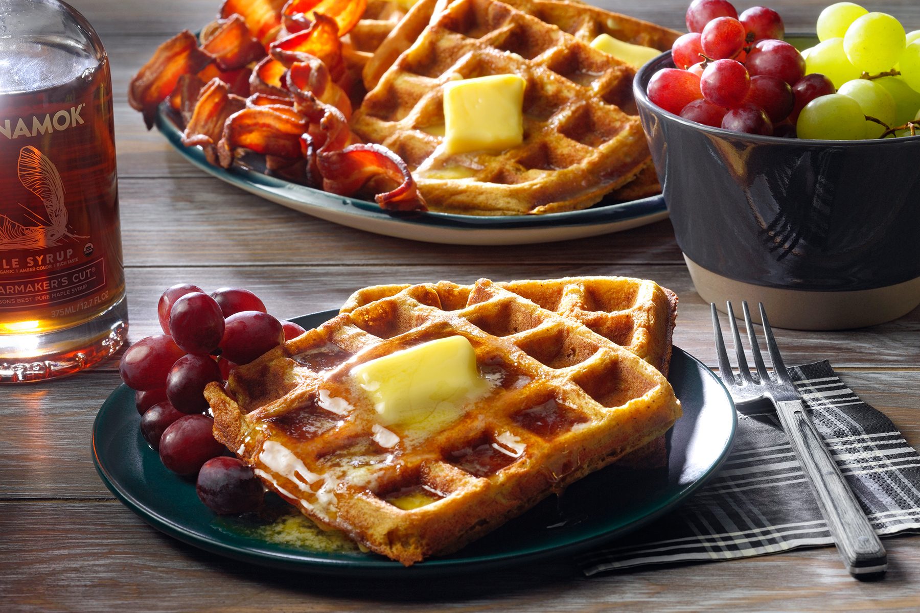 A breakfast spread featuring waffles topped with butter and syrup, accompanied by crispy bacon strips, and a small bunch of red and green grapes. A bottle of maple syrup sits at the side. Forks and a napkin are positioned next to the plates.