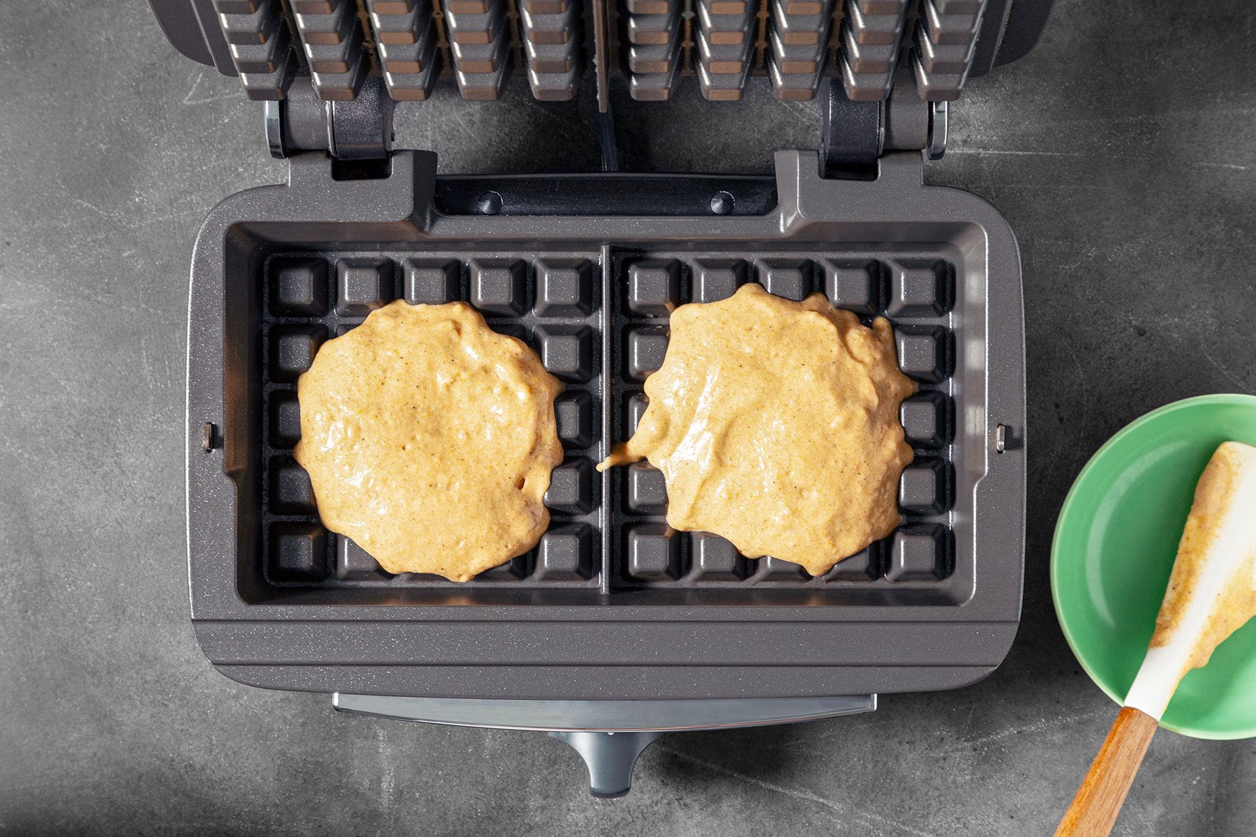 An open waffle maker with two dollops of batter on the grids, ready to be cooked. A green bowl with a spatula rests beside the appliance on a dark countertop.