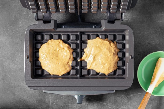 An open waffle maker with two dollops of batter on the grids, ready to be cooked. A green bowl with a spatula rests beside the appliance on a dark countertop.