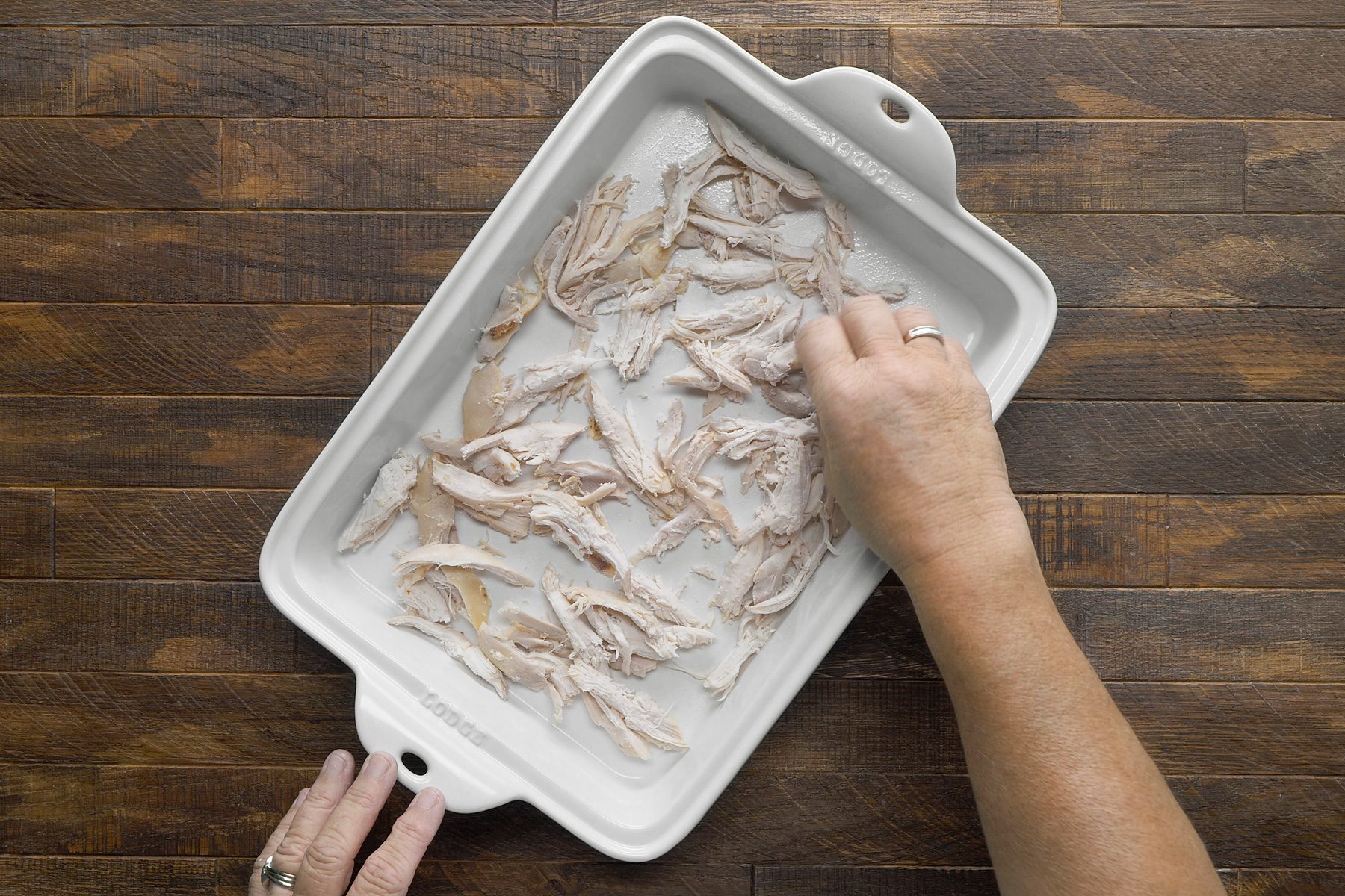overhead shot; wooden background; A white baking dish contains shredded chicken, A person's hand is sprinkling a white powder, likely flour, over the chicken, The chicken is pale white and the flour is covering the chicken evenly