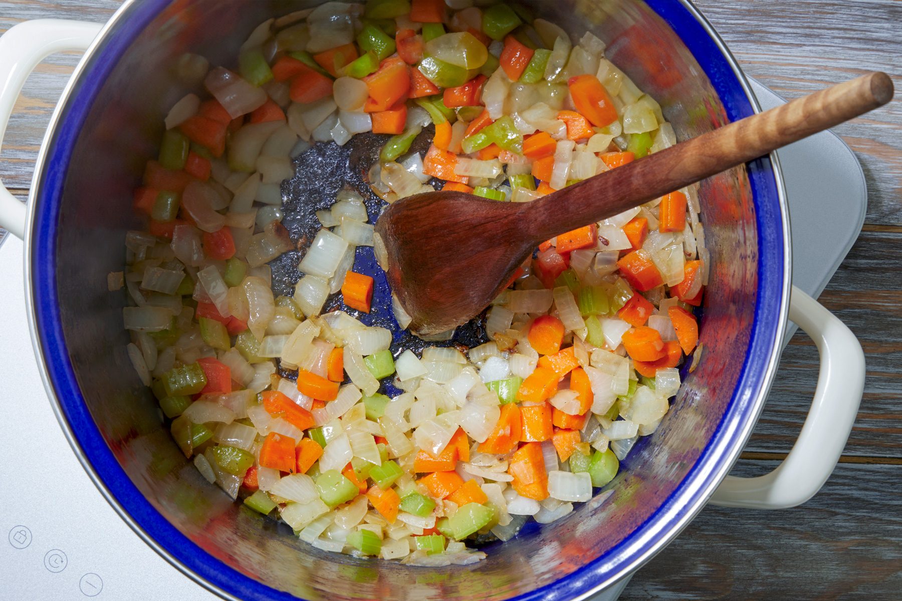 overhead shot; wooden background; Added onion, carrots and celery to same pan and cooking, a wooden spatula can be seen in the pan