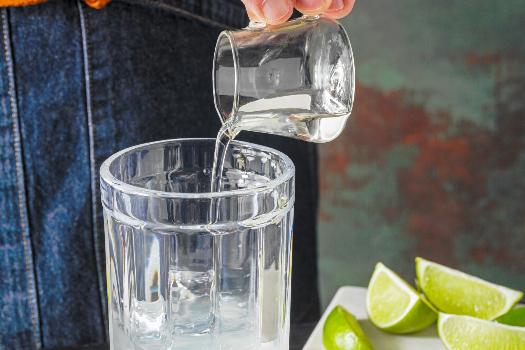 High agnle view shot of adding simple syrup; lime wedges; chopping board; green and brown texture wall; black wooden base;